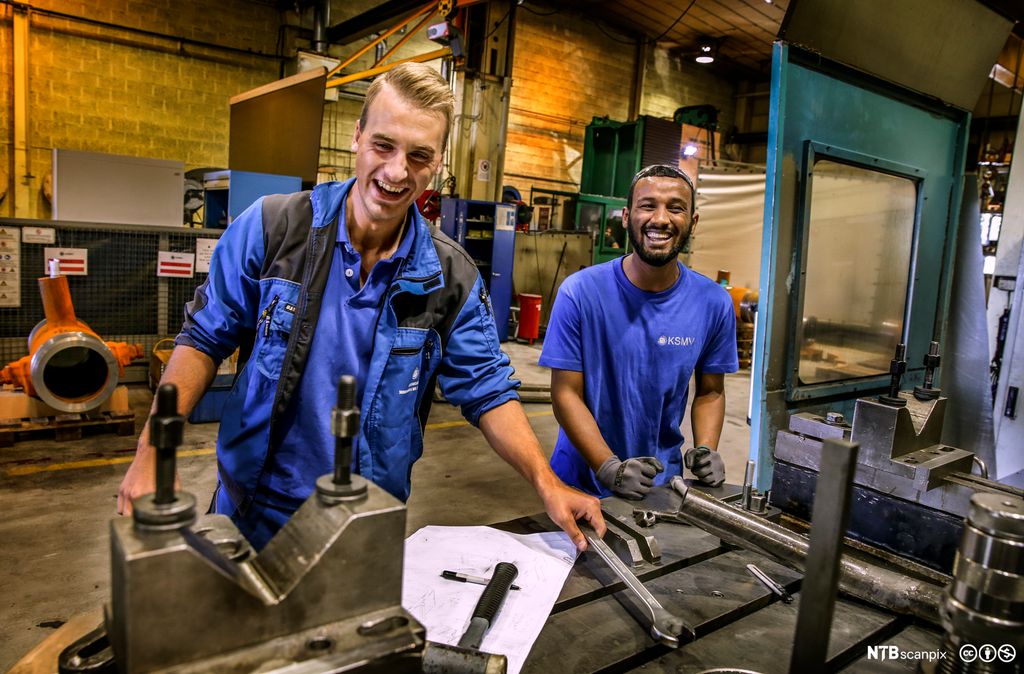 To lærlinger på et mekanisk verksted. De står ved arbeidsbenken og smiler bredt. Foto.