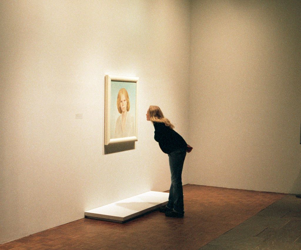 A young woman in an art museum is studying a portrait of another woman. She's bending forwards, studying the details of the painting. Photo.