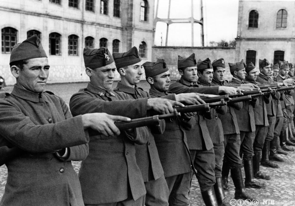 Ei lang rekke uniformskledde menn står oppstilt med geværer rettet framover. Foto. 