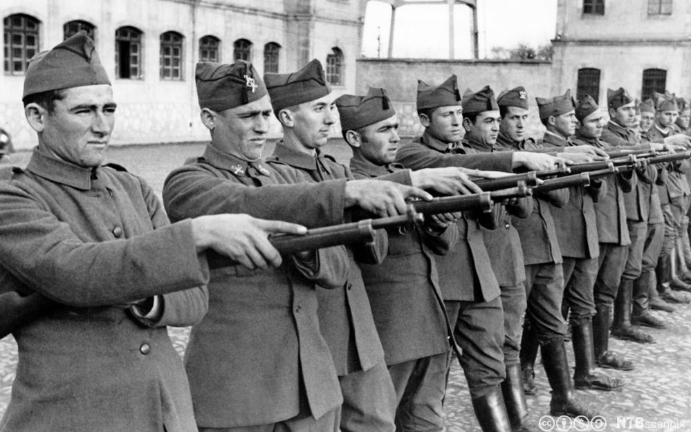 Ei lang rekke uniformskledde menn står oppstilt med geværer rettet framover. Foto.