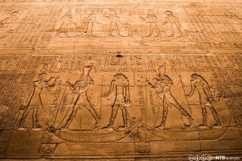 Inngravert utsmykning i egyptisk tempel som vi blant annet viser mennesker stående i båter. Foto. 
