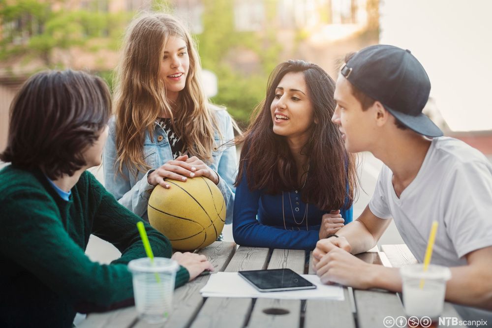 Fire ungdommar sit rundt eit bord i skulegarden og pratar. Foto.