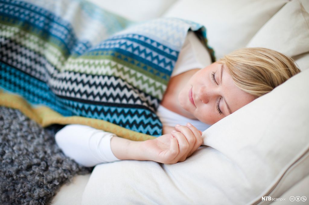 Ung kvinne ligger på en lys sofa og sover. Hun har et mønstra pledd over seg. Foto.