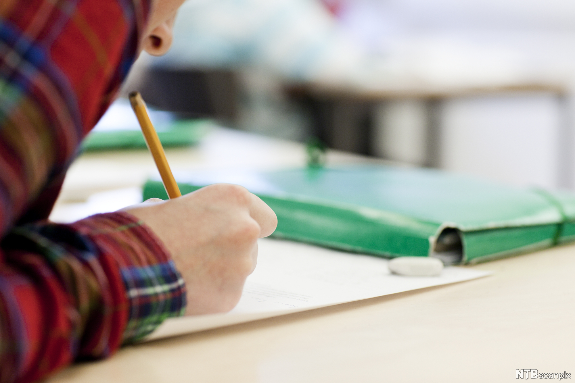 A person in a red shirt is writing. They are using a yellow pencil. In front of them on the desk is a green folder. Photo.