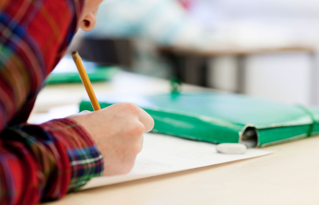 A person in a red shirt is writing. They are using a yellow pencil. In front of them on the desk is a green folder. Photo.