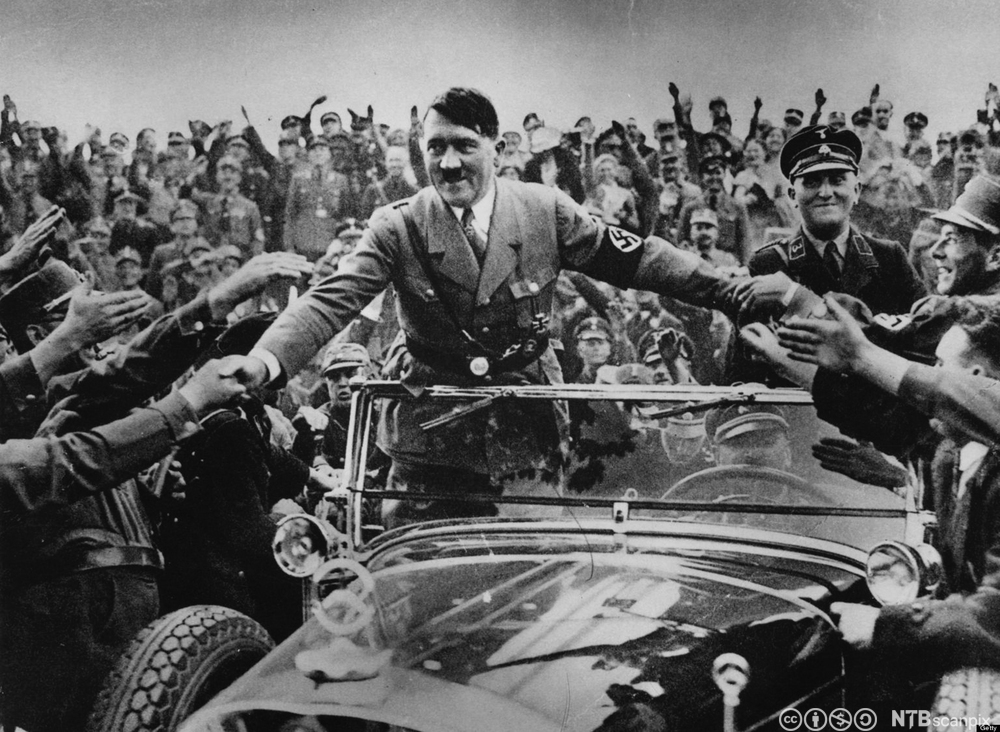 Hitler i uniform og med armbind med hakekors står i en åpen bil og hilser på tilhengere langs veien. Foto. 