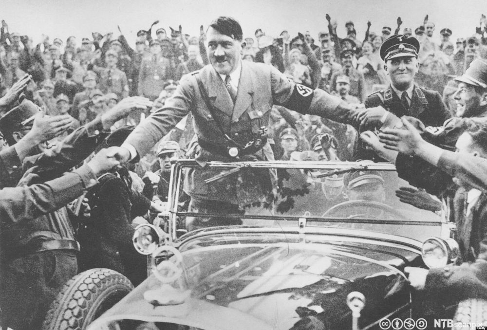 Hitler i uniform og med armbind med hakekors står i en åpen bil og hilser på tilhengere langs veien. Foto.