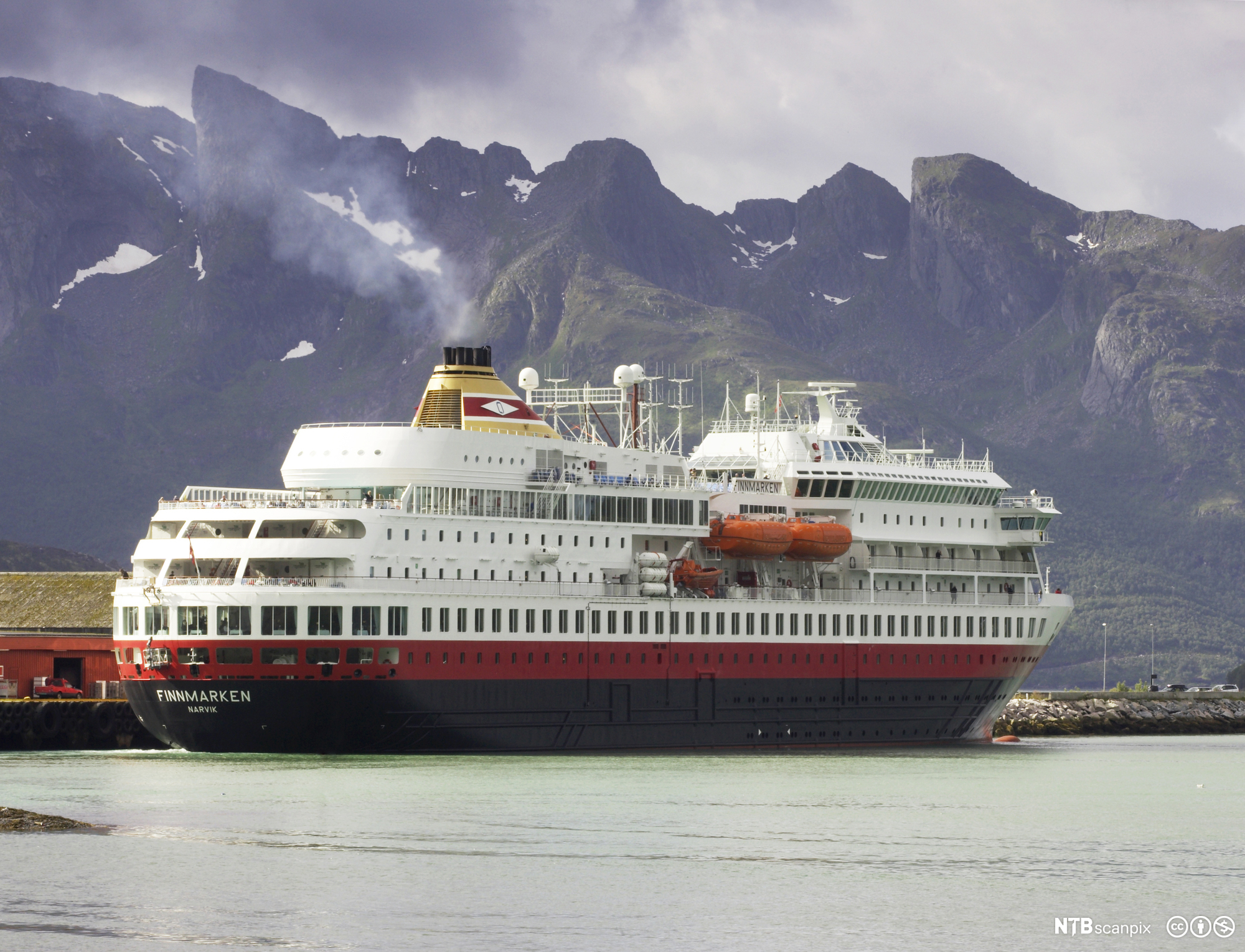 Stort passasjerskip med navnet Finnmarken ved kai. I bakgrunnen stiger høye, skogkledte fjell mot himmelen. Det er sommer, men enkelte snøflekker ligger igjen mot toppen. Foto.