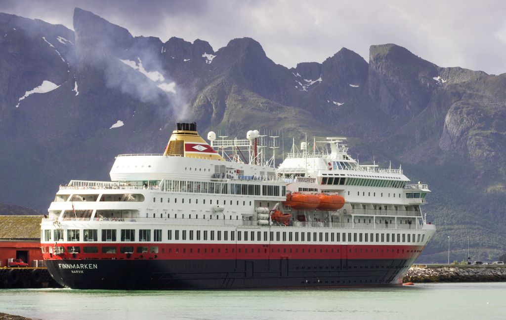 Stort passasjerskip med navnet Finnmarken ved kai. I bakgrunnen stiger høye, skogkledte fjell mot himmelen. Det er sommer, men enkelte snøflekker ligger igjen mot toppen. Foto.