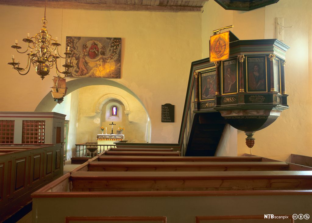 Interiør frå kvitmåla steinkyrkje. Kyrkjebenker, altar og prekestol. Foto.