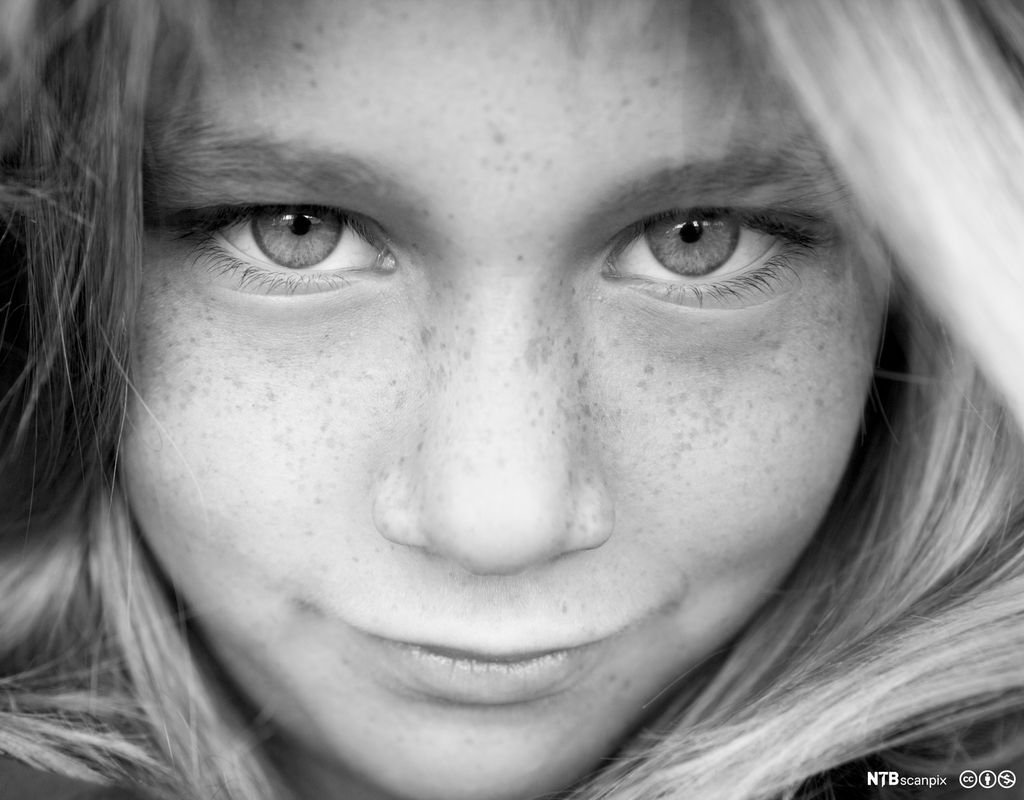 Ansiktet på ei jente med lyst hår, lyse øyne og fregner over nesa. Hun smiler litt og ser rett på betrakteren. Foto.