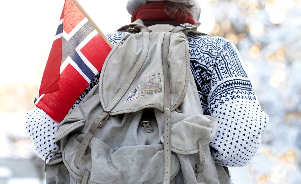 Utsnitt av person i lusekofte sett bakfra, med norsk flagg stikkende opp av en gammeldags ryggsekk. Foto.