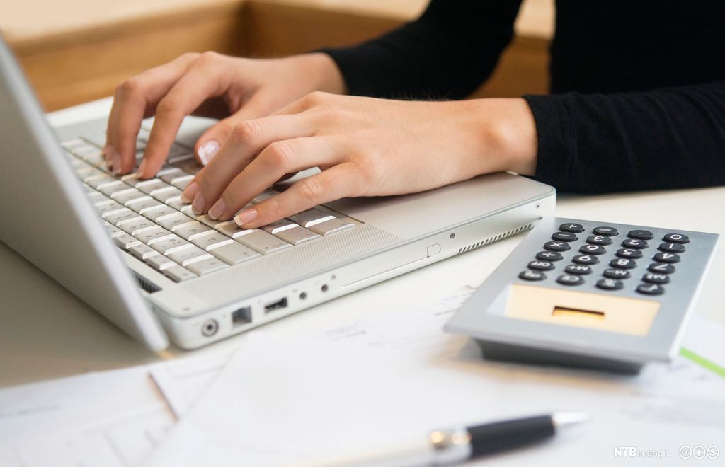 En person skriver på en laptop, ved siden av ligger det en kalkulator, en penn og noen ark. Foto.