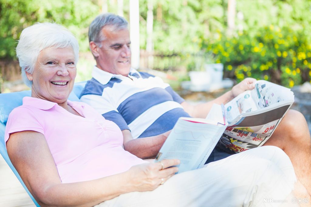 En eldre dame og en eldre mann sitter i solen og leser i henholdsvis en bok og en reklameavis. Foto.