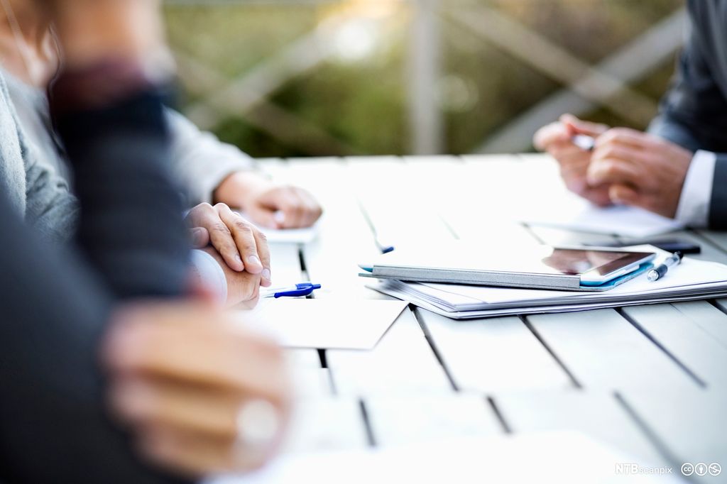 Personer rundt et bord med notater og nettbrett foran seg. Bildet viser bare hendene og bordet. Foto. 