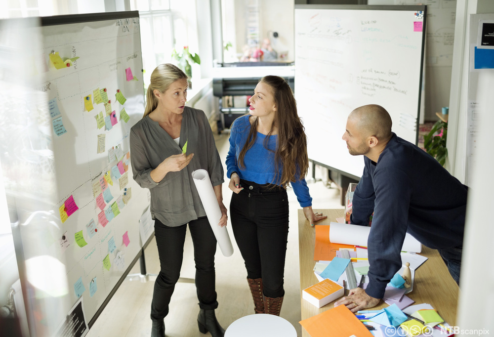 To kvinner og en mann står og diskuterer foran en stor tusjtavle i et kontorlokale. På tavlen henger det påskrevne post-it-lapper i ulike farger. Foto.