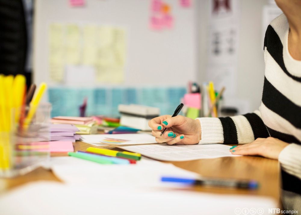 Kvinne sit ved eit bord og skriv for hand. Fotografiet viser berre hendene, den eine armen og litt av overkroppen hennar. Ark, pennar og blokker med post-it-lappar ligg utover bordet. Foto.