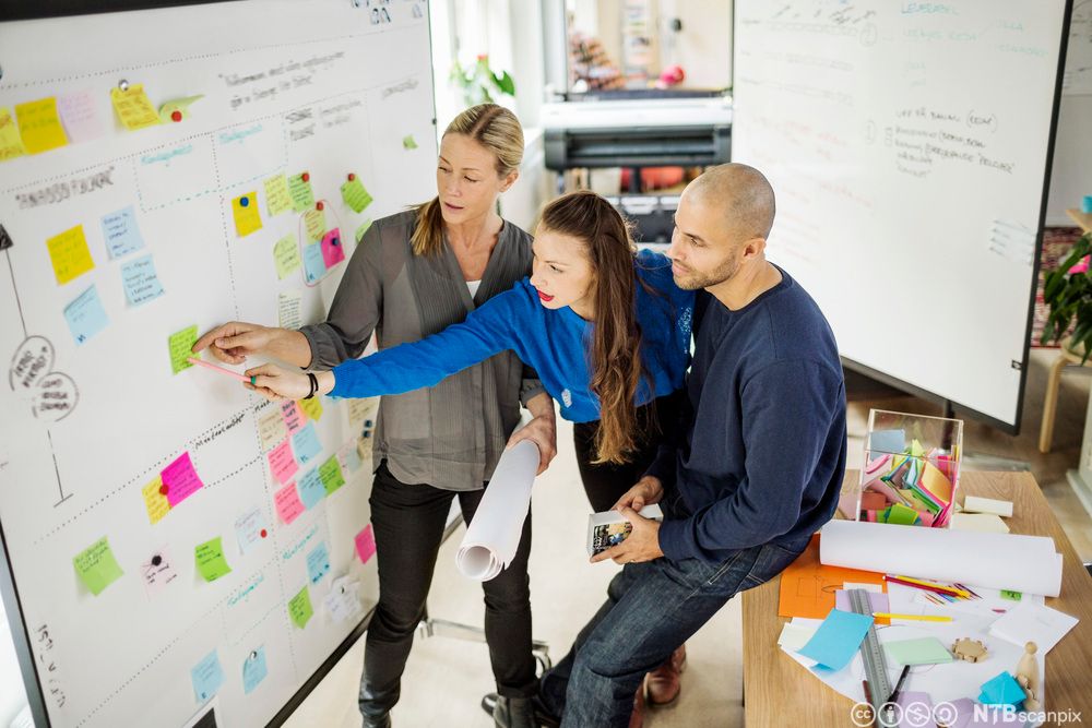 Forretningsfolk diskuterer idéer på et kontor. De har ei tavle foran seg med post-it-lapper i ulike farger. To av personene peker på en av post-it-lappene, og en annen sitter på en pult og følger med. Foto.