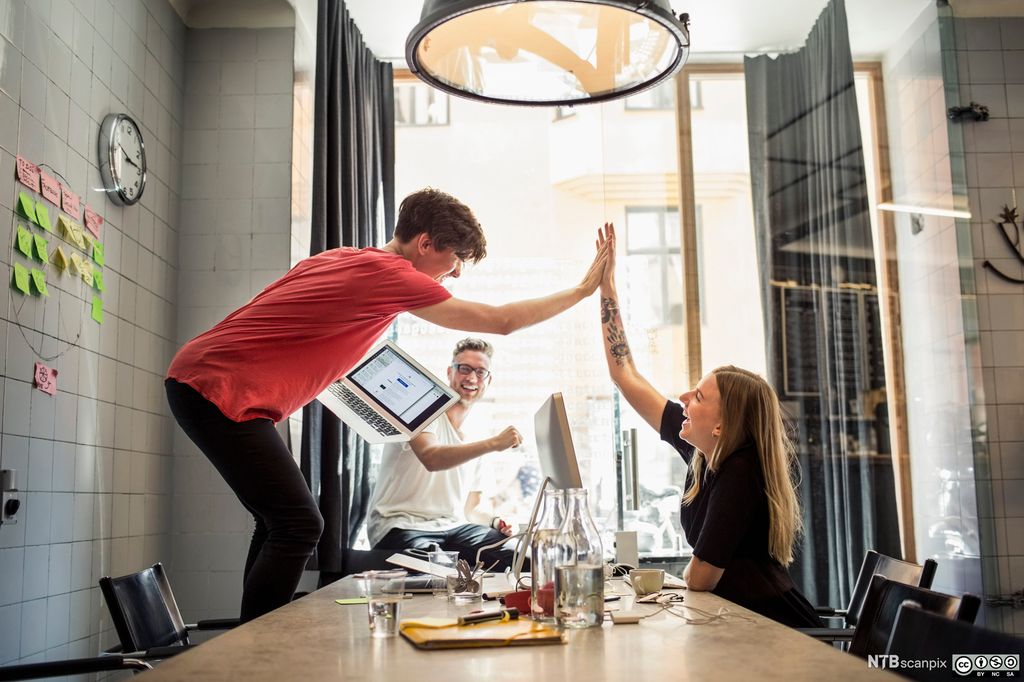 To gründere jobber ved et stort bord. De gir en "high five" til hverandre. Den ene av dem holder en pc i den ene hånden. Den tredje gründeren i rommet sitter i vinduskarmen og gir sin fulle støtte ved hjelp av et stort smil og positivt kroppsspråk. Det står vannflasker, kopper og glass p bordet. Det ligger også skrivesaker der, og en pc er plassert i midten.  På en av veggene er det klistret opp post-it-lapper i ulike farger. Foto.