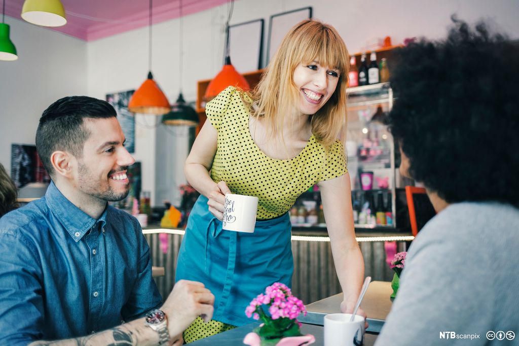Smilande servitør set drikke på bordet til to gjester på kafé. Foto.