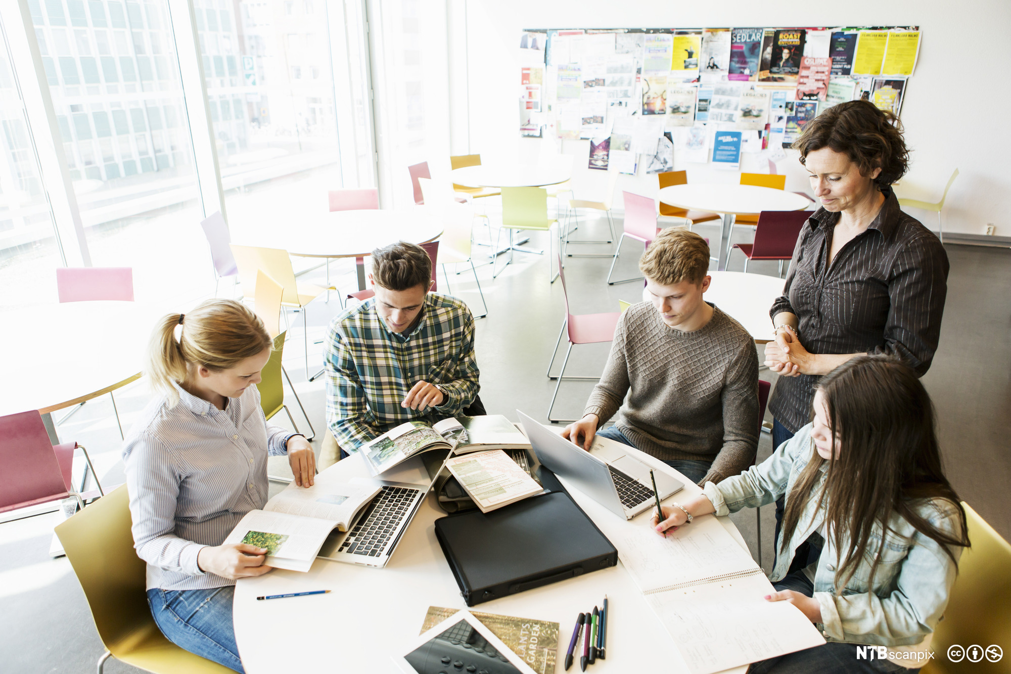 Lærer og elever i klasserom. Elevene sitter ved et rundt bord og blar i bøker, skriver og leser på pc. Læreren står ved siden av og observerer dem. Foto.