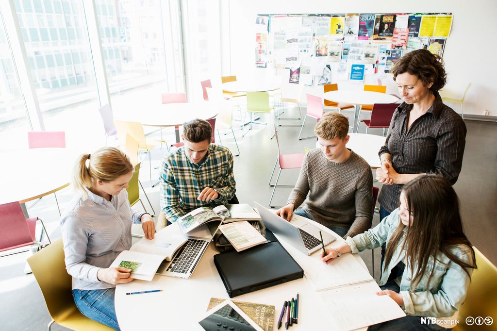 Fire ungdommer sitter ved et rundt bord og jobber med bøker og datamaskiner. En voksen person står ved siden av og ser på. Foto.