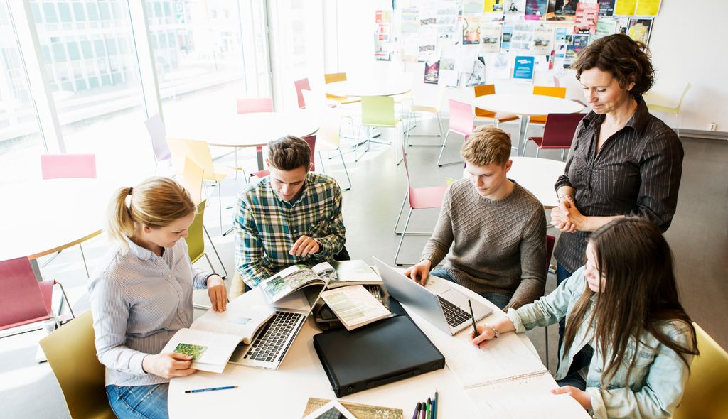 Lærer og elever i klasserom. Fire elever sitter rundt et skrivebord. Læreren står og observerer dem. Foto.