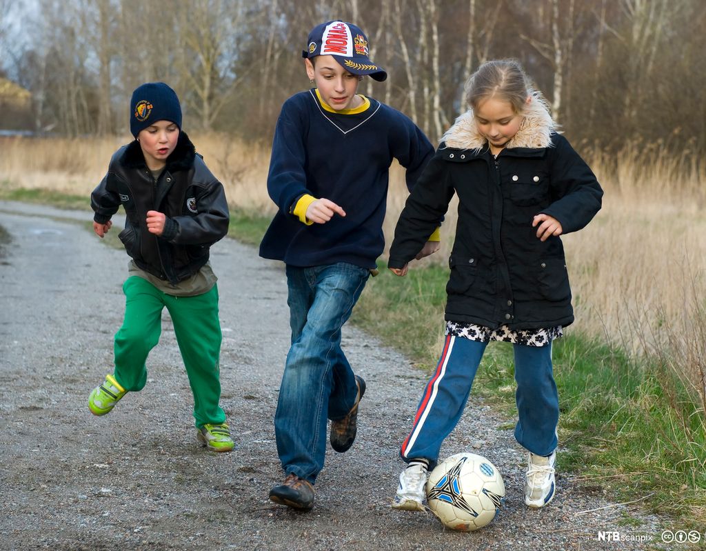 Tre barn som leker med en fotball på en grusvei. Foto.