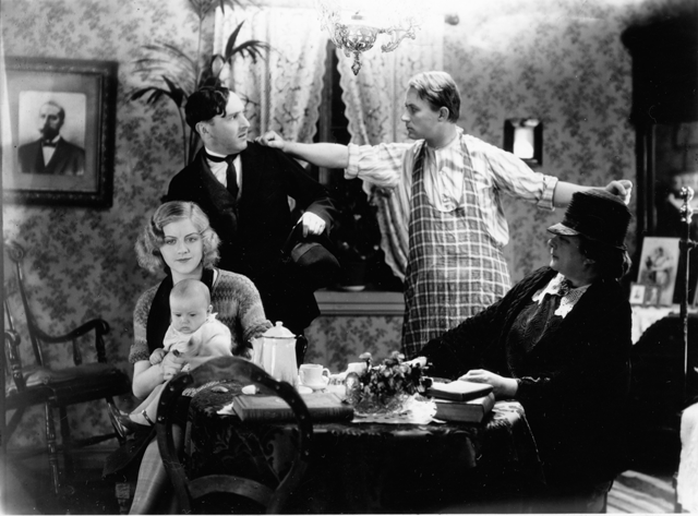 En ung og en eldre kvinne sitter ved et bord. Den unge kvinner har en baby på fanget. Bak står to menn som krangler. Foto.