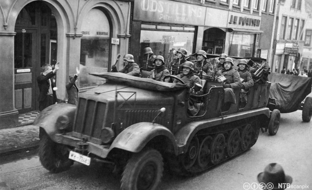 Mange tyske soldater i en åpen beltebil kjører inn i Aabenraa i Danmark, 9. april 1940. På fortauet har et par tilskuere hevet høyre hånd i en mulig nazihilsen. Foto. 