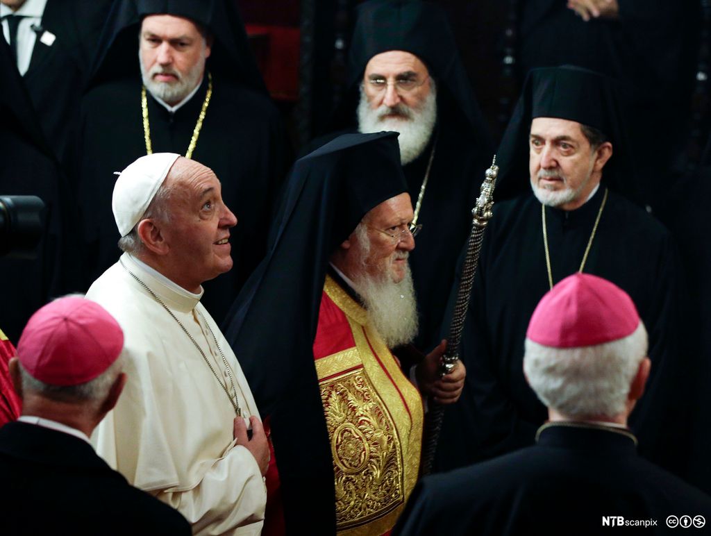 Pave Frans og pariark Bartholomeus I av Konstantinopel går ved siden av hverandre. Paven har en hvit kappe og hvitt hodeplagg. Patriarken har en overdel i rødt og gull med sort kappe over og sort hodeplagg. De er omgitt av menn med sorte kapper. Foto.