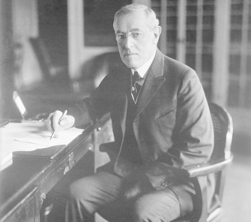 President Woodrow Wilson sitter ved skrivebordet. Han har en penn i høyre hånd, og noen papirer ligger foran ham. Han ser mot fotografen. Foto.