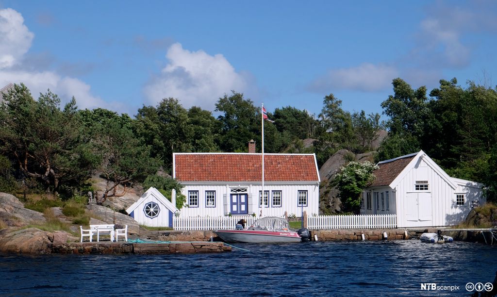 Hvitt sørlandshus med bå inngangsdør ved sjøen. Foto.