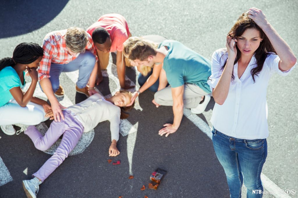 Ung kvinne ringer etter hjelp ved en ulykke. En person ligger på bakken bak henne, flere andre hjelper den skadde personen. Foto.