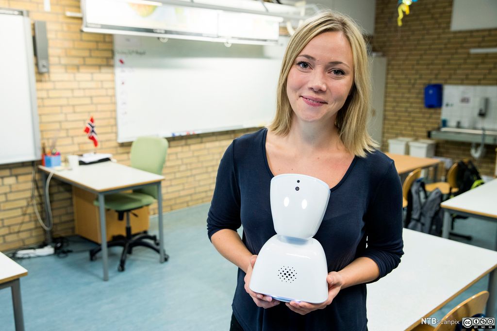 Smilende kvinne holder en liten robot i et klasserom. Roboten er hvit. Den er todelt og den øverste delen ligner et hode med lysende øyne. Foran på den nedre delen er det en høyttaler. Foto.