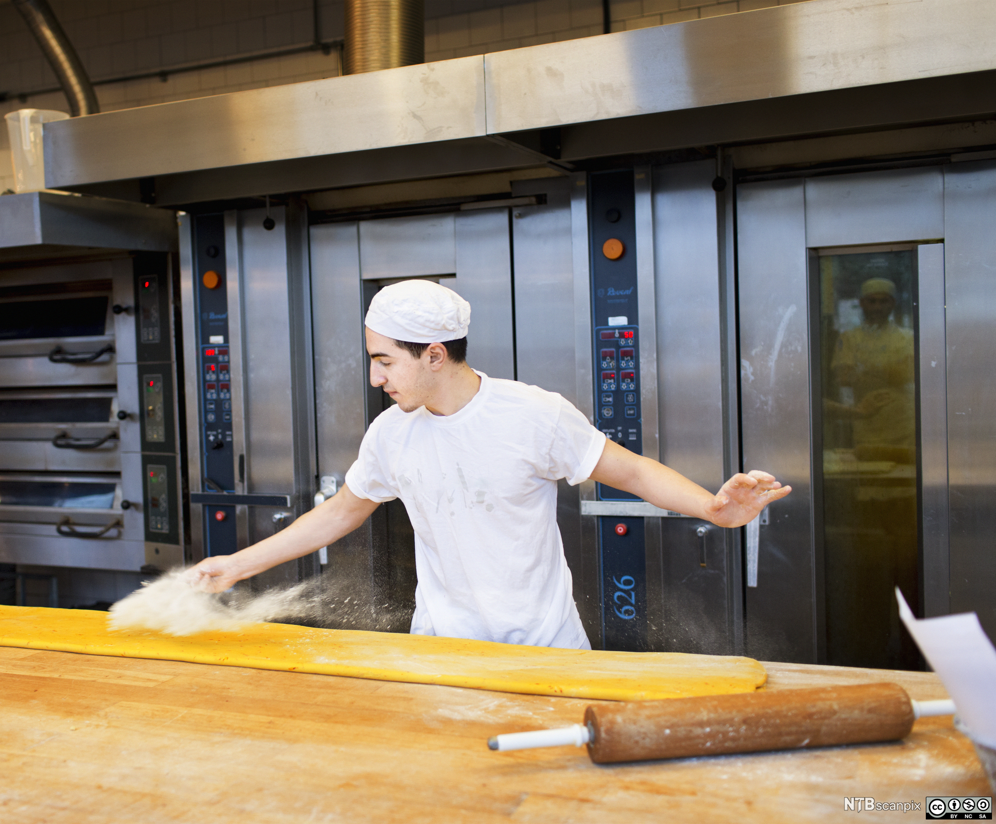 En baker strør mel over en utrullet deig på et bord. Foto.