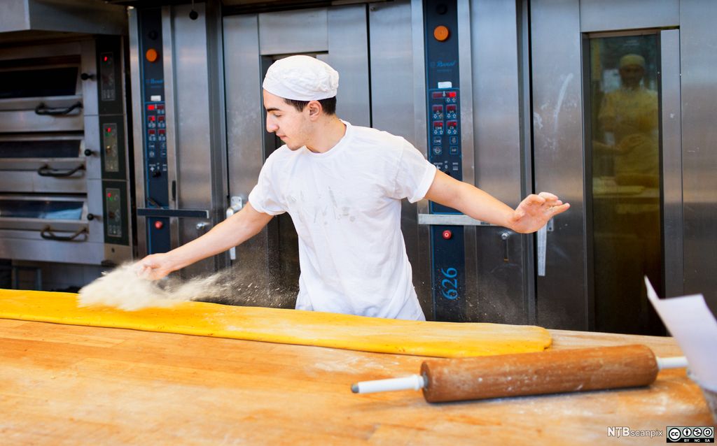 En baker strør mel over en utrullet deig på et bord. Foto.