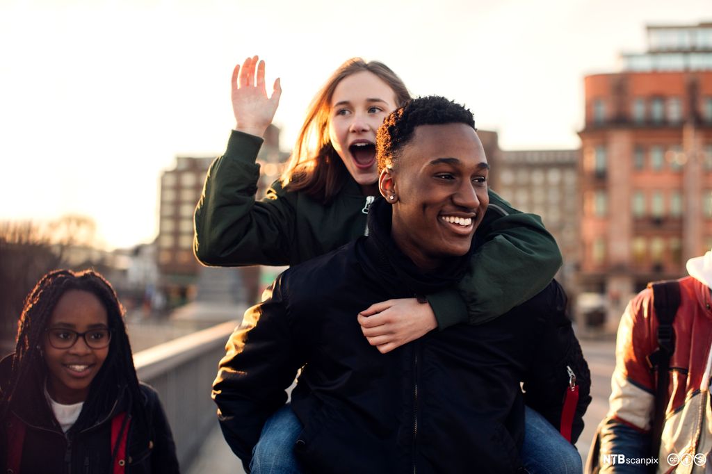 Ei ung jente sitter på ryggen til en ung gutt. Begge ler. Ei annen smilende jente går sammen med dem i gata. Foto.