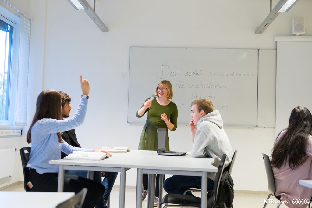 Elevar og lærarar i klasserom på vidaregåande skule. Læraren står framme ved tavla. Elevane sit gruppevis ved pultar, og éin elev rekker opp handa. Foto.