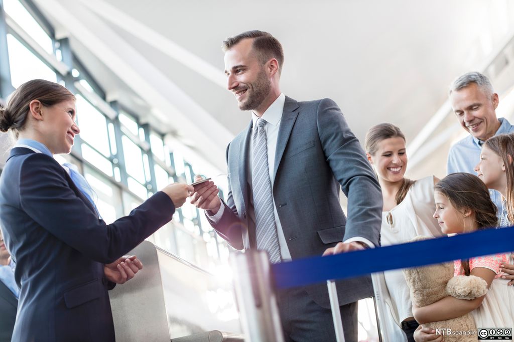 Servicemedarbeidar med uniform på flyplass. Ho smiler til ein passasjer med trillekoffert. Foto. 