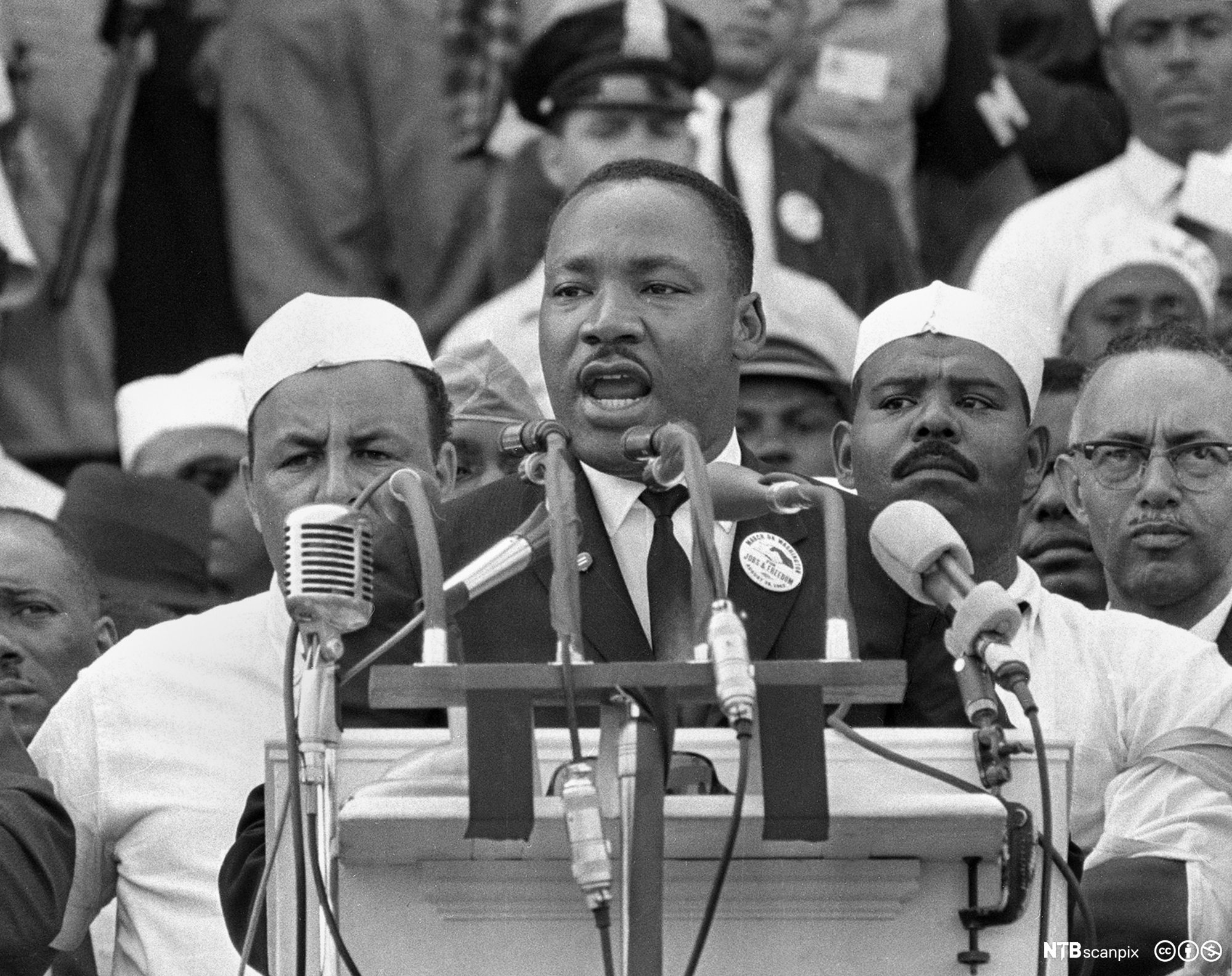Photo: We see a Black man standing behind a lectern. Many microphones are attached to the lectern. Behind him we see several people. 