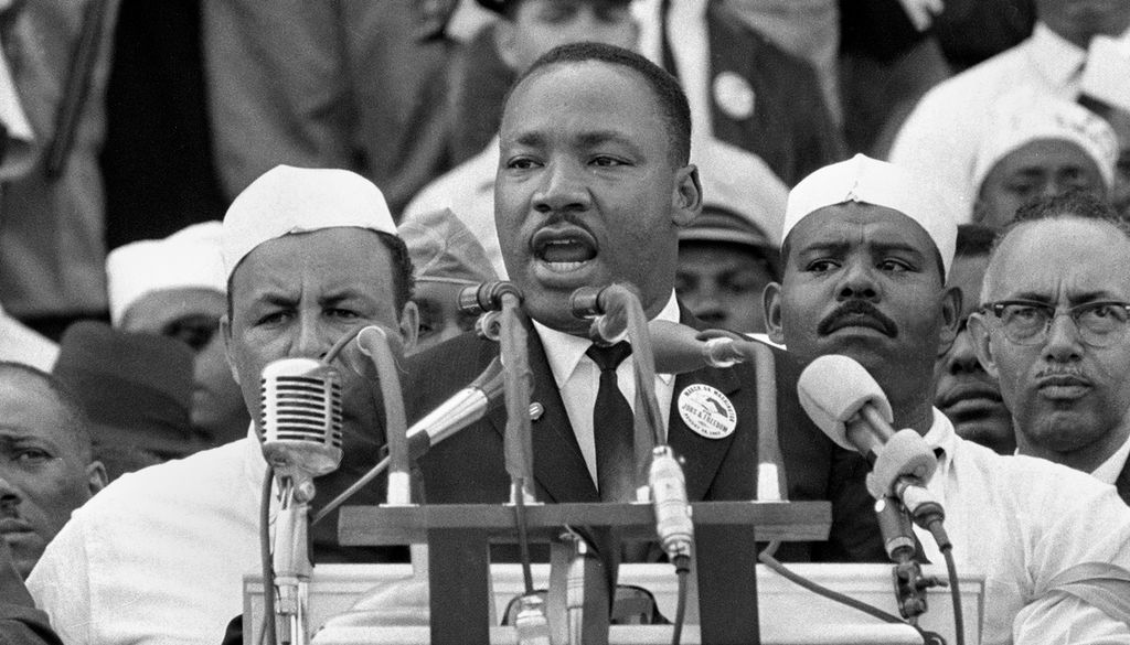 Photo: We see a Black man standing behind a lectern. Many microphones are attached to the lectern. Behind him we see several people.
