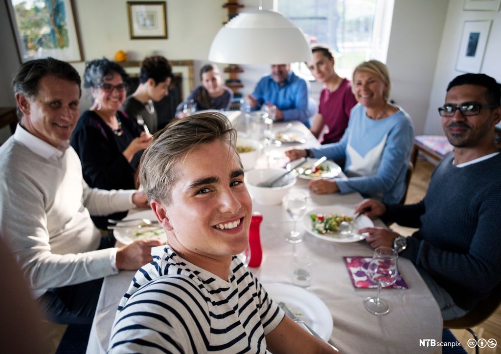 Ein tenåringsgut saman med familie og venner rundt eit middagsbord. Foto.