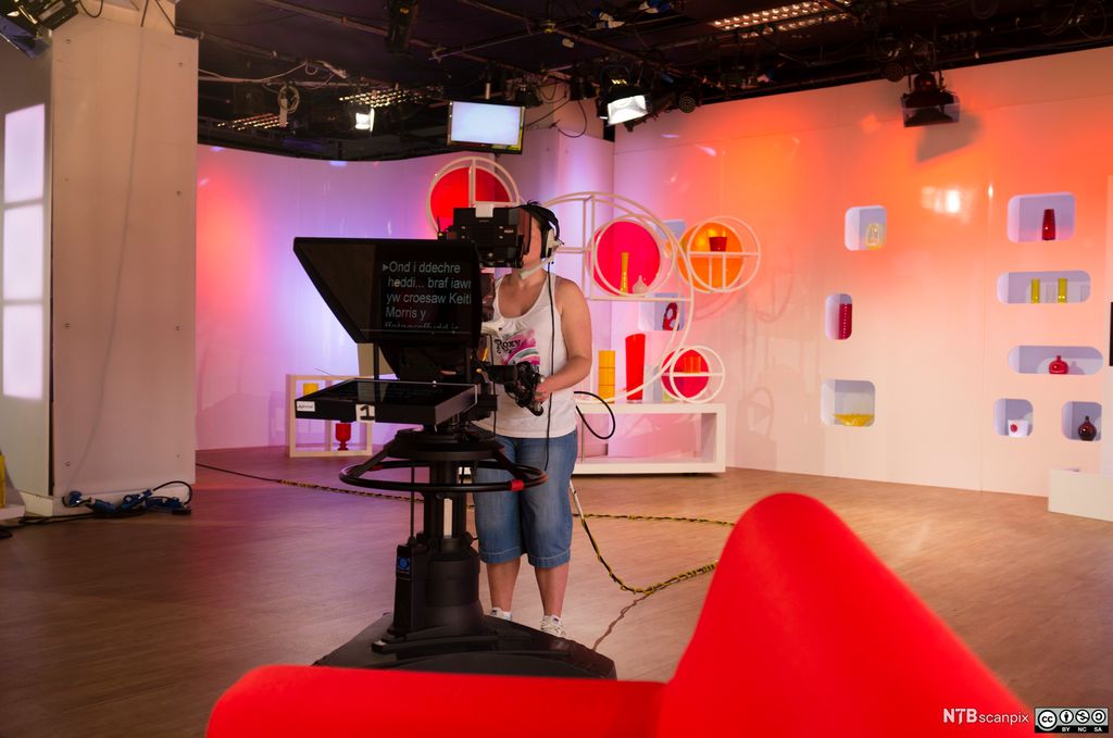 Ei dame står bak et TV-kamera i et fargerikt TV-studio. Foto.