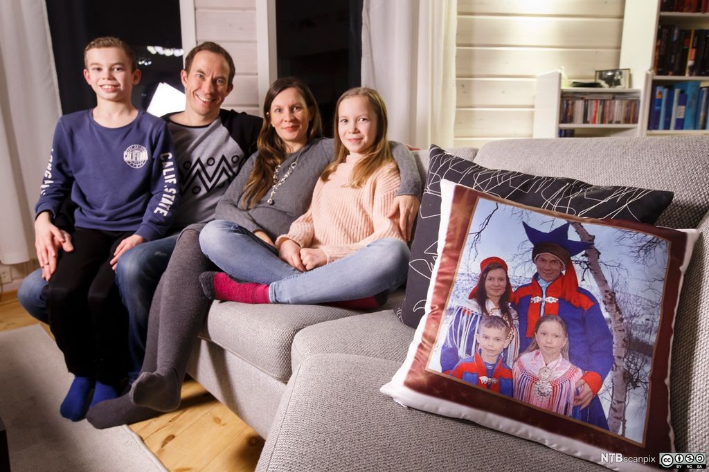 En mann, en kvinne, en gutt og ei jente sitter i en sofa. I sofaen er det også ei pute med bilde av de samme personene i samekofter. Foto.