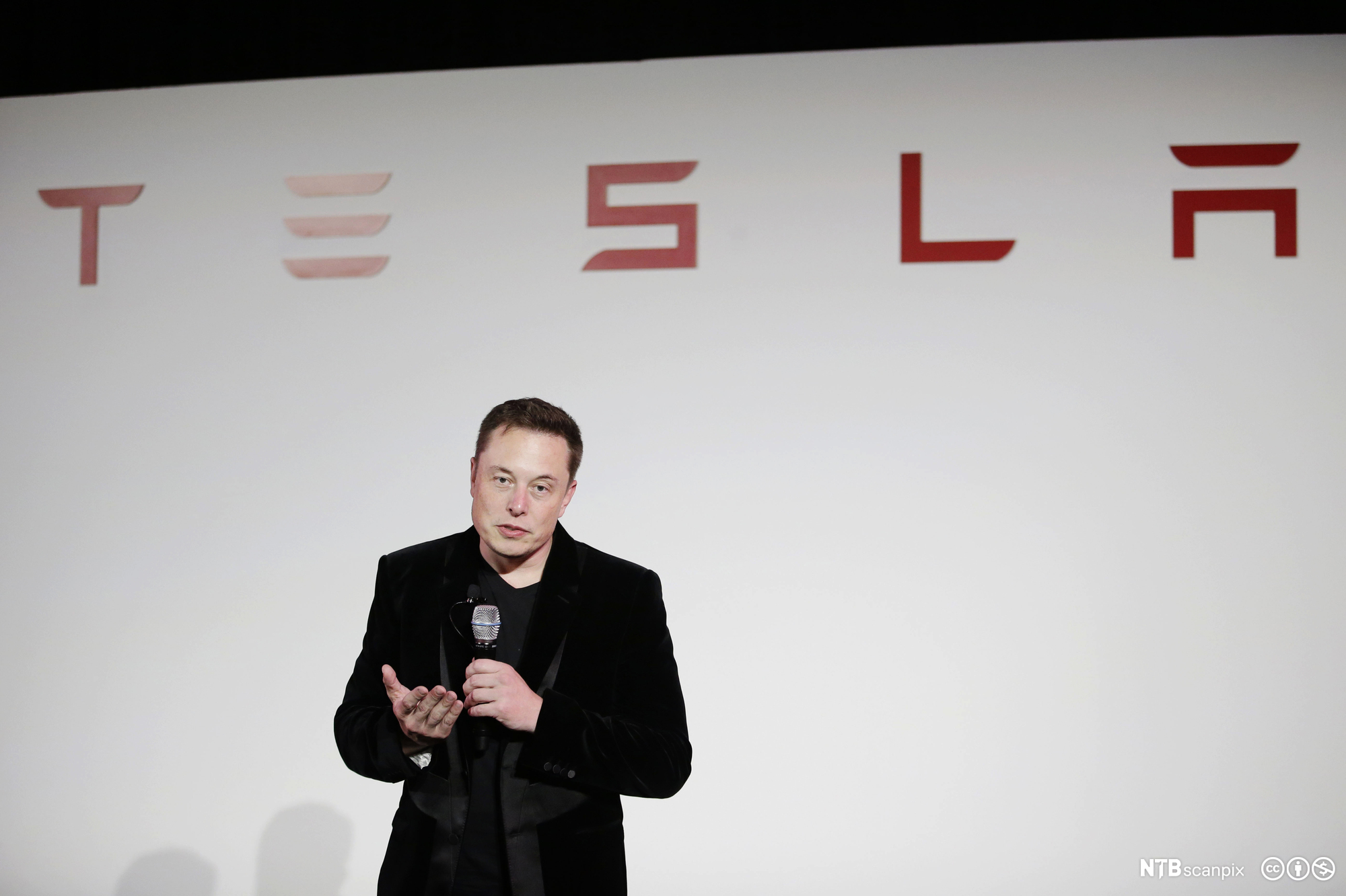 Elon Musk prater i mikofon under en Tesla-logo. Foto.