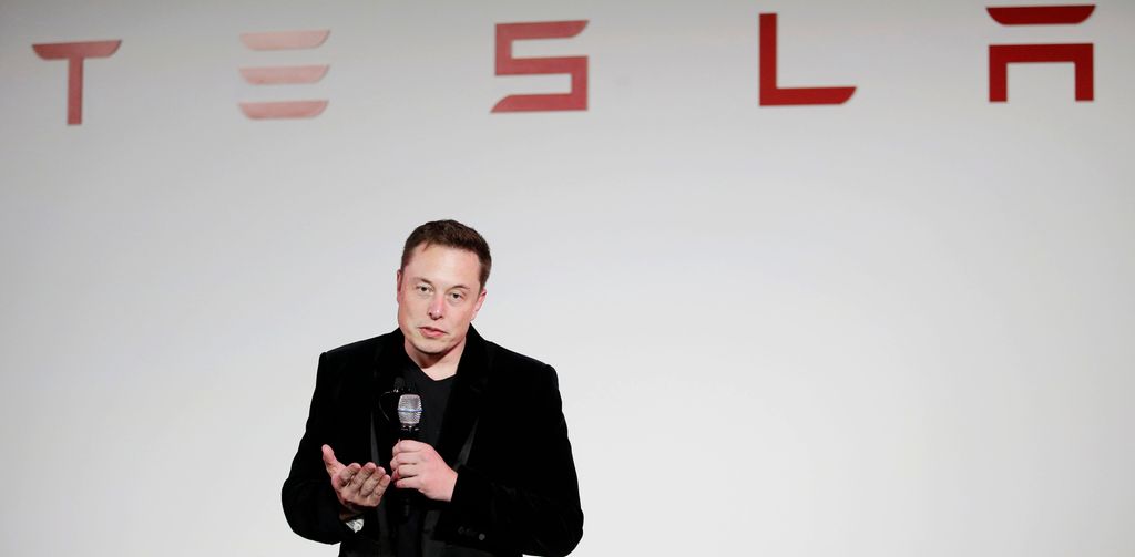 Elon Musk prater i mikofon under en Tesla-logo. Foto.