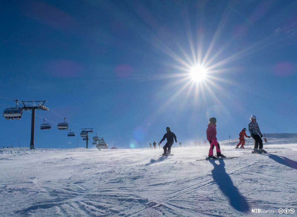 Folk på ski og snøbrett i ein slalåmbakke på ein fin dag med strålande sol. Foto.