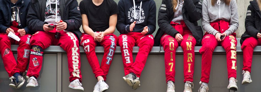 En rekke avgangselever sitter på en lang benk. Bildet er beskåret slik at vi ser russebuksene deres, som er røde og dekorert med store hvite og gullfargede bokstaver og ulike strykemerker. Foto