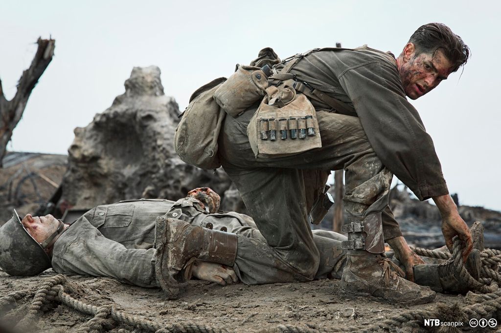 Soldat sitter på huk på en slagmark. Bak ham ligger en død eller skadet soldat. Foto.